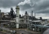 Willis Unveils Carbon Capture and Storage Insurance Solution Industrial chemical plant with a tall smokestack emitting white vapor under dark, cloudy skies, surrounded by interconnected pipes and metal structures.