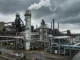 Willis Unveils Carbon Capture and Storage Insurance Solution Industrial chemical plant with a tall smokestack emitting white vapor under dark, cloudy skies, surrounded by interconnected pipes and metal structures.