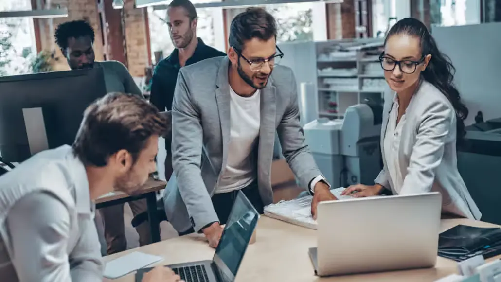 Team of professionals collaborating around a desk with laptops in a modern office, discussing a project.