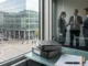 Europe Banking Sector May Raise Lending by €2tn Office desk by a window overlooking a city street; three professionals converse behind a glass partition, partially visible through the window reflection, with binders and papers on the desk.