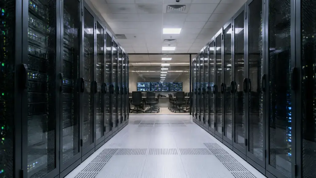 Data center corridor flanked by glass-front server racks, leading to a control room with chairs and multiple screens at the far end.