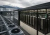 Blue Owl Leads $2.4 Billion AI Factory Financing for IREN Row of outdoor cooling units beside a data center server racks housed in a glass-enclosed enclosure, with mountains in the distance.