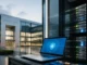 Munich Re Plans Cyber Insurance Acquisition of At-Bay Laptop showing a blue security shield screen beside a row of server racks outside a modern glass office building.